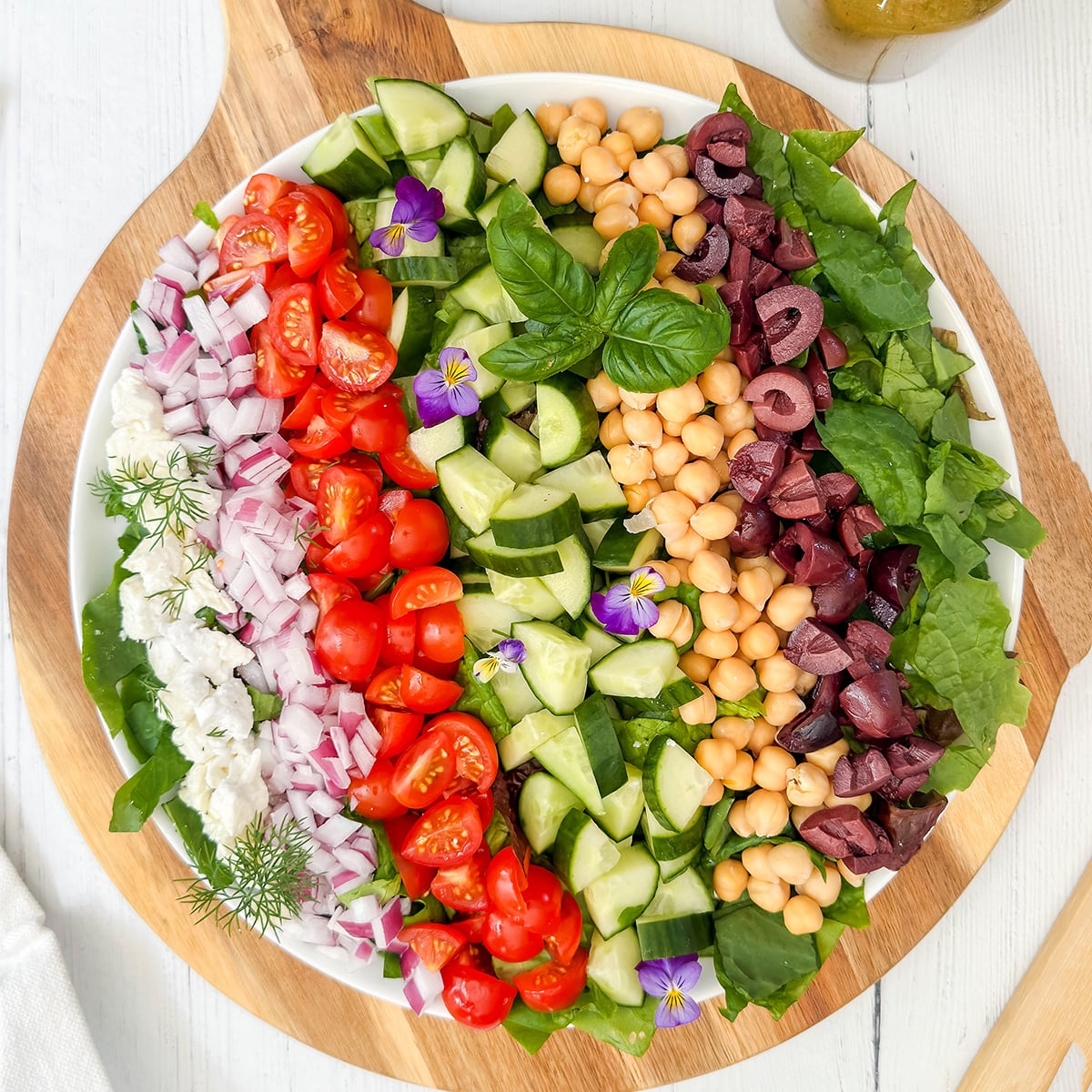 A large round platter with rows of chopped tomatoes, cucumbers, red onions, olives, chickpeas, feta cheese, and spinach—this stunning display celebrates the vibrant flavors of Mediterranean salads, garnished with fresh herbs and edible flowers.