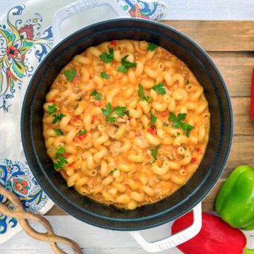 A pot of creamy taco pasta with ground meat, chopped red peppers, and parsley garnish, placed on a wooden surface with bell peppers and a patterned cloth nearby.