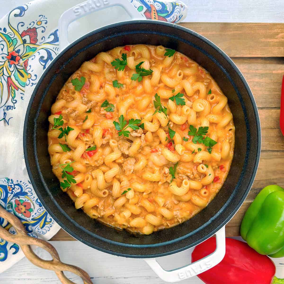 A pot of creamy taco pasta with ground meat, chopped red peppers, and parsley garnish, placed on a wooden surface with bell peppers and a patterned cloth nearby.