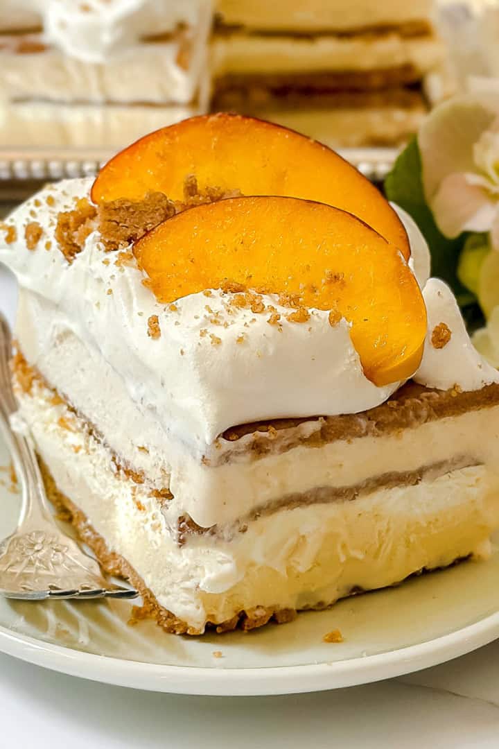 A slice of easy icebox cake layered with whipped cream, graham cracker crumbs, and peach slices sits on a white plate with a fork.