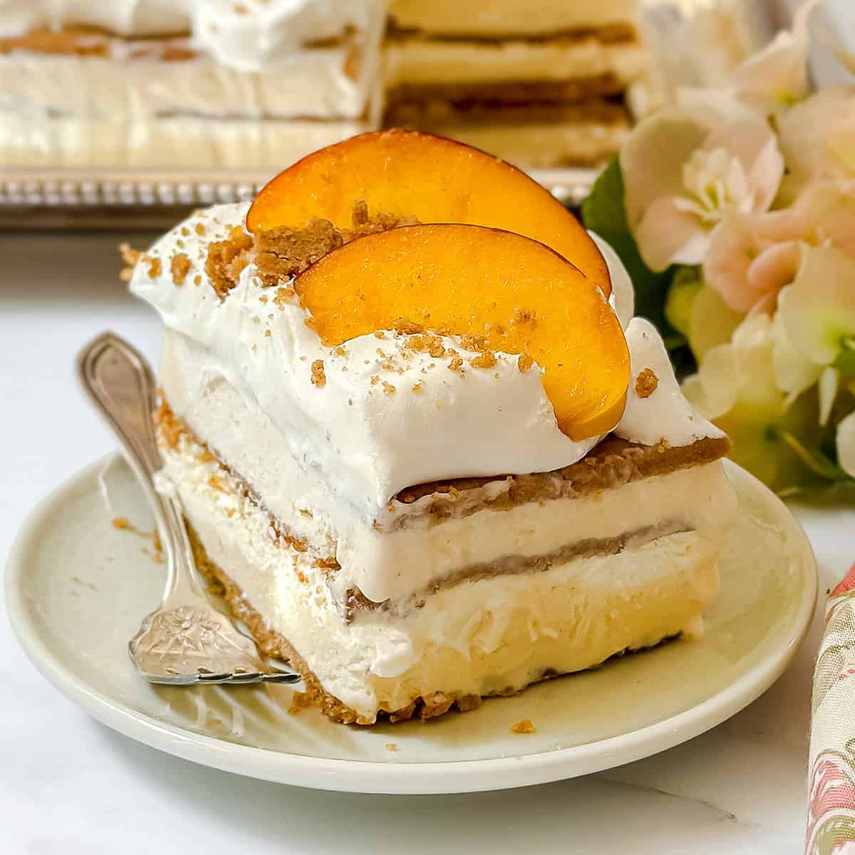 A slice of easy icebox cake layered with whipped cream, graham cracker crumbs, and peach slices sits on a white plate with a fork.