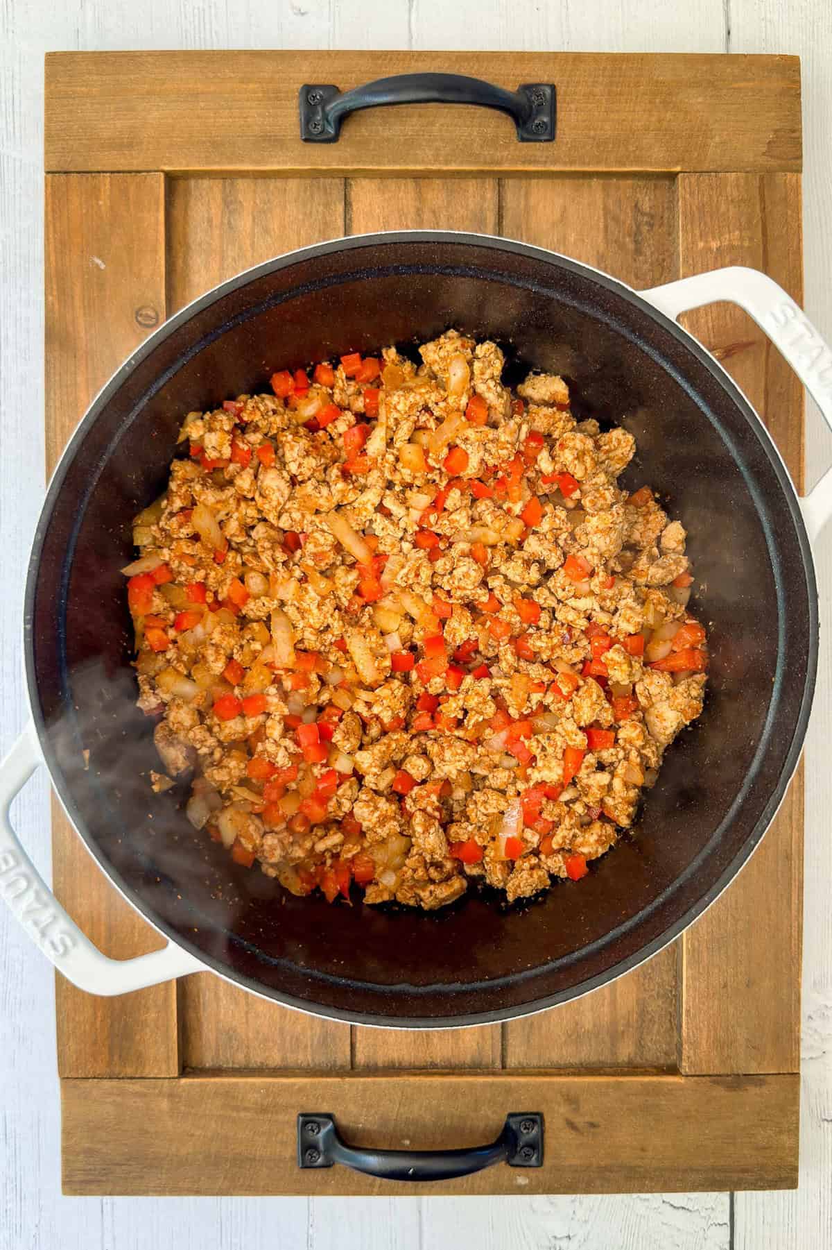 A white pot on a wooden surface filled with browning ground meat, diced red bell peppers, and onions.