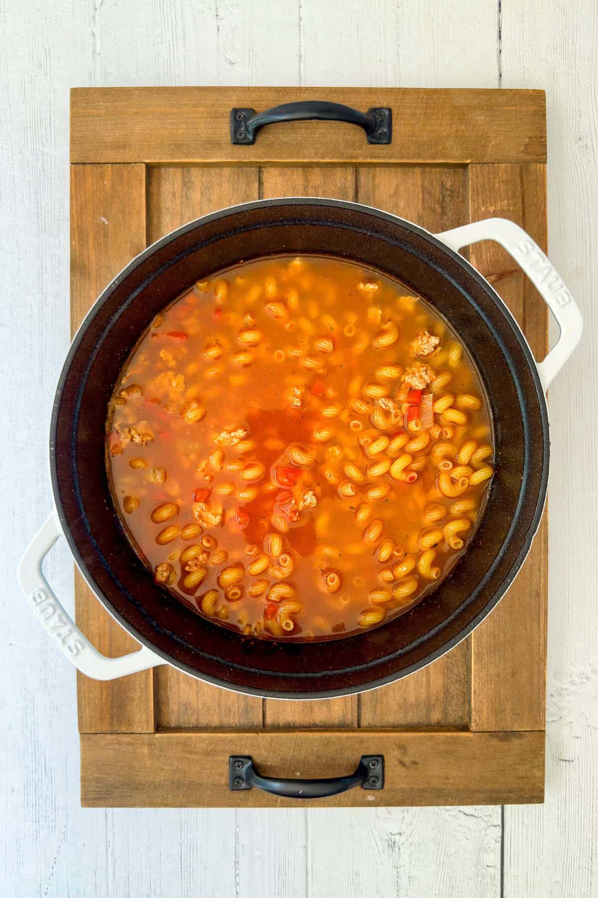 A pot of taco pasta soup with ground meat, pasta, and tomatoes sits on a wooden board atop a white table.