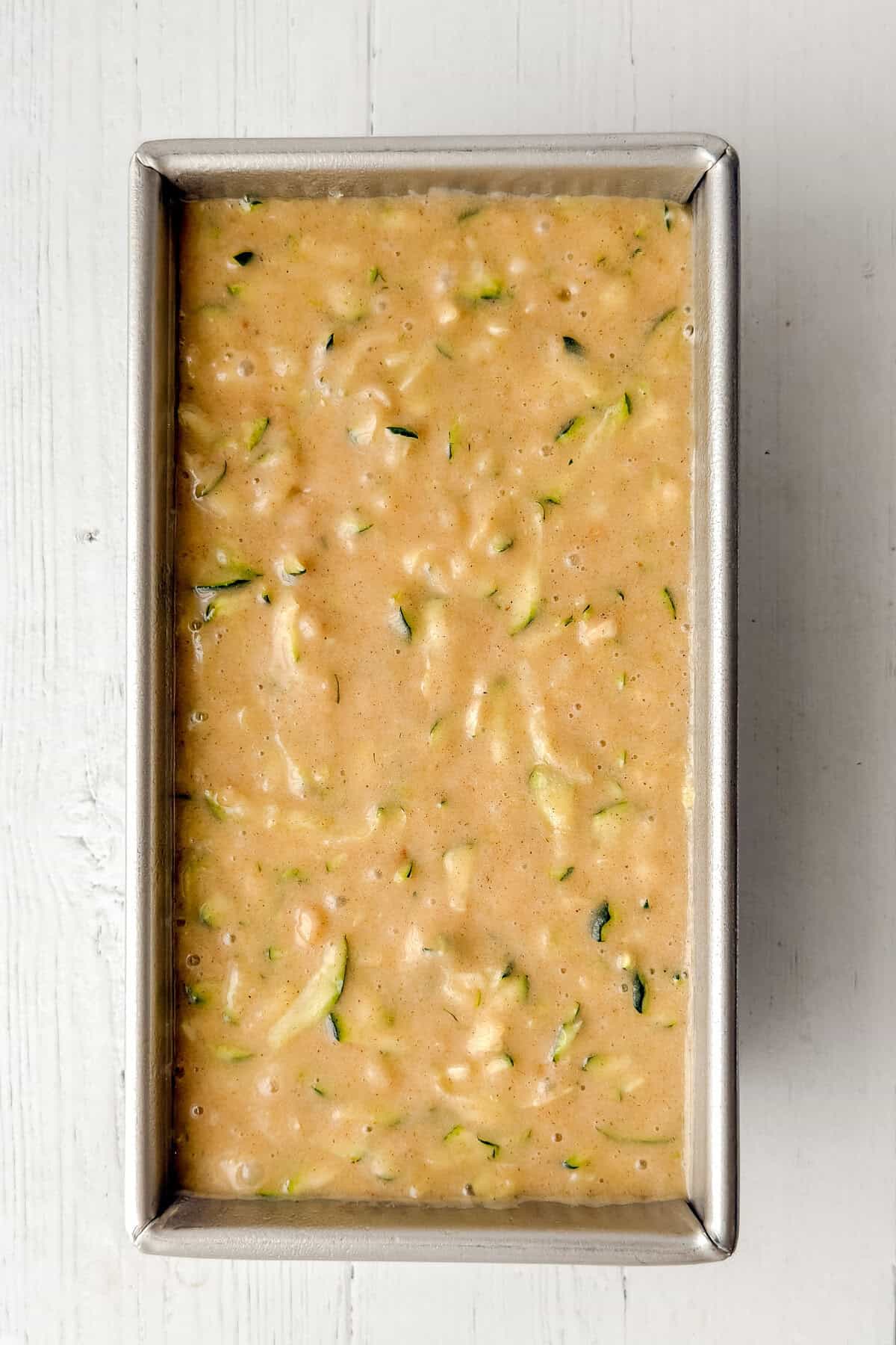 A metal loaf pan filled with raw Zucchini Bread batter sits on a white wooden surface, showing visible bits of shredded zucchini throughout the light tan mixture.