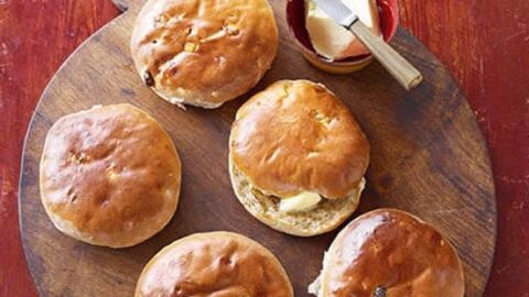 A round wooden cutting board holds five freshly baked bread rolls. There is a small red bowl with pats of butter and a butter knife resting on the side of the board. The background is a rustic, textured red surface.