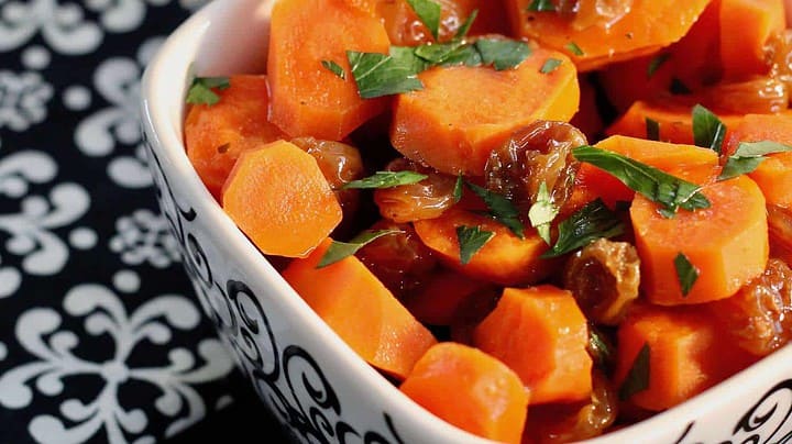 A bowl of glazed carrots garnished with chopped parsley. The carrots are sliced and mixed with raisins, creating a vibrant and colorful dish. The white bowl with a black pattern sits on a black and white patterned tablecloth.