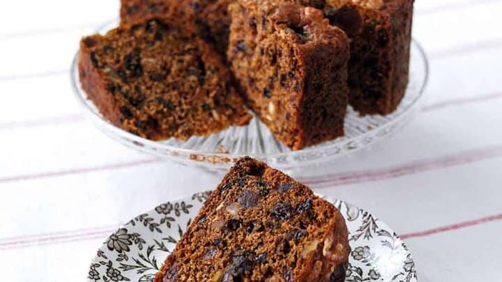 A rich fruit cake sits on a glass stand, with a few slices cut and one slice placed on a floral-patterned plate in the foreground. The cake is dense, filled with assorted nuts and dried fruits, and a white tablecloth with red stripes lies underneath.