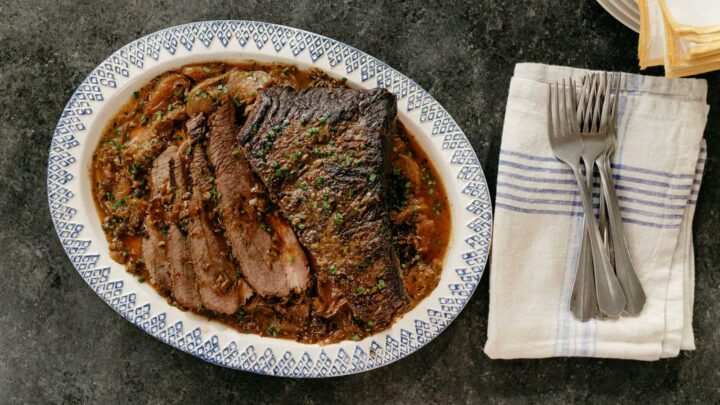 A serving plate of sliced roast beef covered in gravy, garnished with fresh herbs, is set on a dark surface. Next to it, a stack of white plates with a yellow rim and a folded white cloth with blue stripes are accompanied by a set of forks.