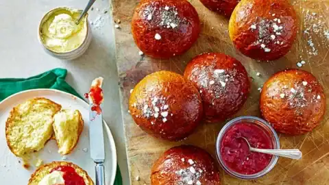 A wooden board with seven golden-brown buns dusted with powdered sugar. Next to the board, a plate holds a split teacake topped with butter and jam. A jar of red jam with a spoon lies nearby, alongside a cup of tea and a bowl of whipped butter. A green napkin is partially visible.