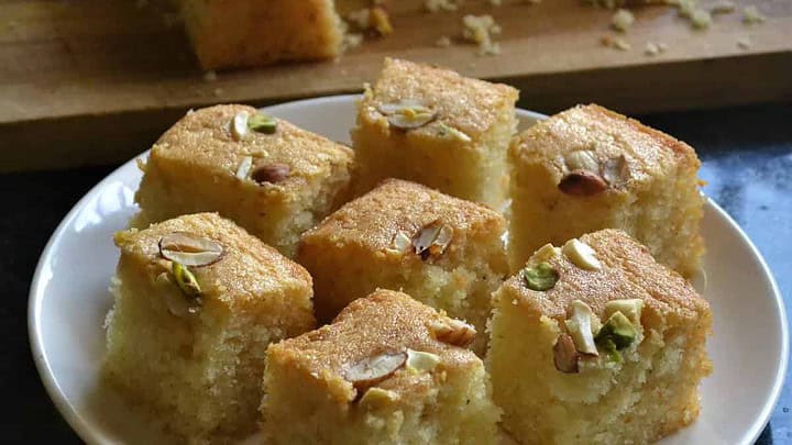A plate of nine square pieces of sponge cake topped with sliced almonds and pistachios. The pieces are golden and moist, placed neatly on a white plate. In the background, more cake is visible on a wooden cutting board.