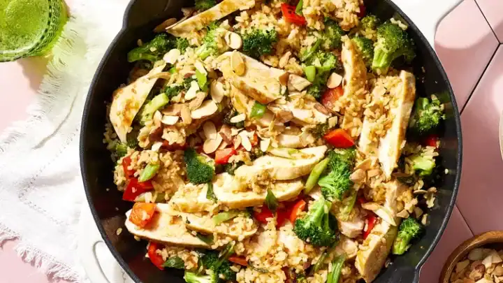 A skillet filled with a vibrant, high-protein chicken and rice dish, garnished with sliced almonds, broccoli, red bell peppers, and seasonings. Two green drinks in glasses sit nearby on a white cloth on a pink hexagonal tiled surface. A small bowl of sliced almonds is also present—a perfect Mediterranean diet dinner.