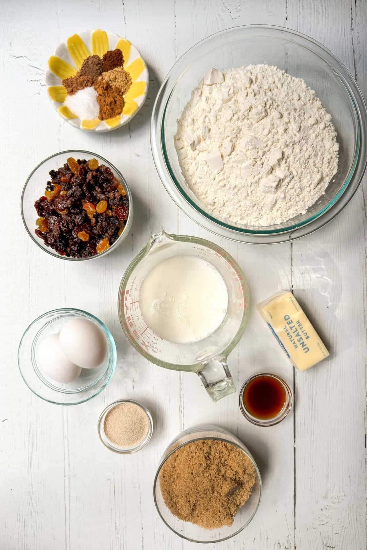 A top-down view of baking ingredients on a white wooden surface. Items include a bowl of flour, a stick of unsalted butter, a small bowl of vanilla extract, two eggs, a bowl of dried fruits, a dish of mixed spices, a measuring cup of milk, and bowls of brown and white sugar.