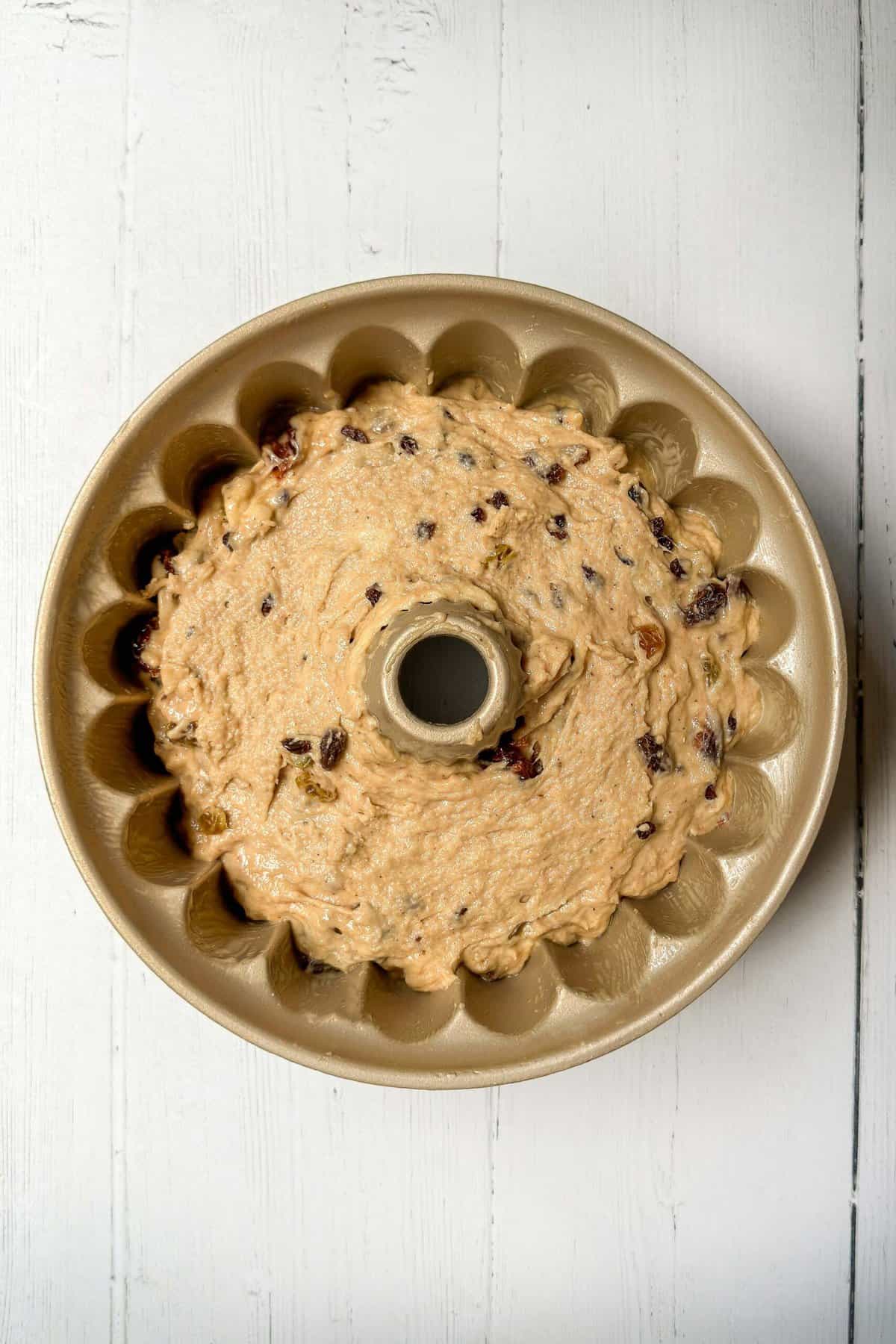 A beige Bundt pan filled with unbaked batter dotted with raisins and other small pieces of fruit, placed on a white wooden surface. The batter appears smooth and ready for baking.