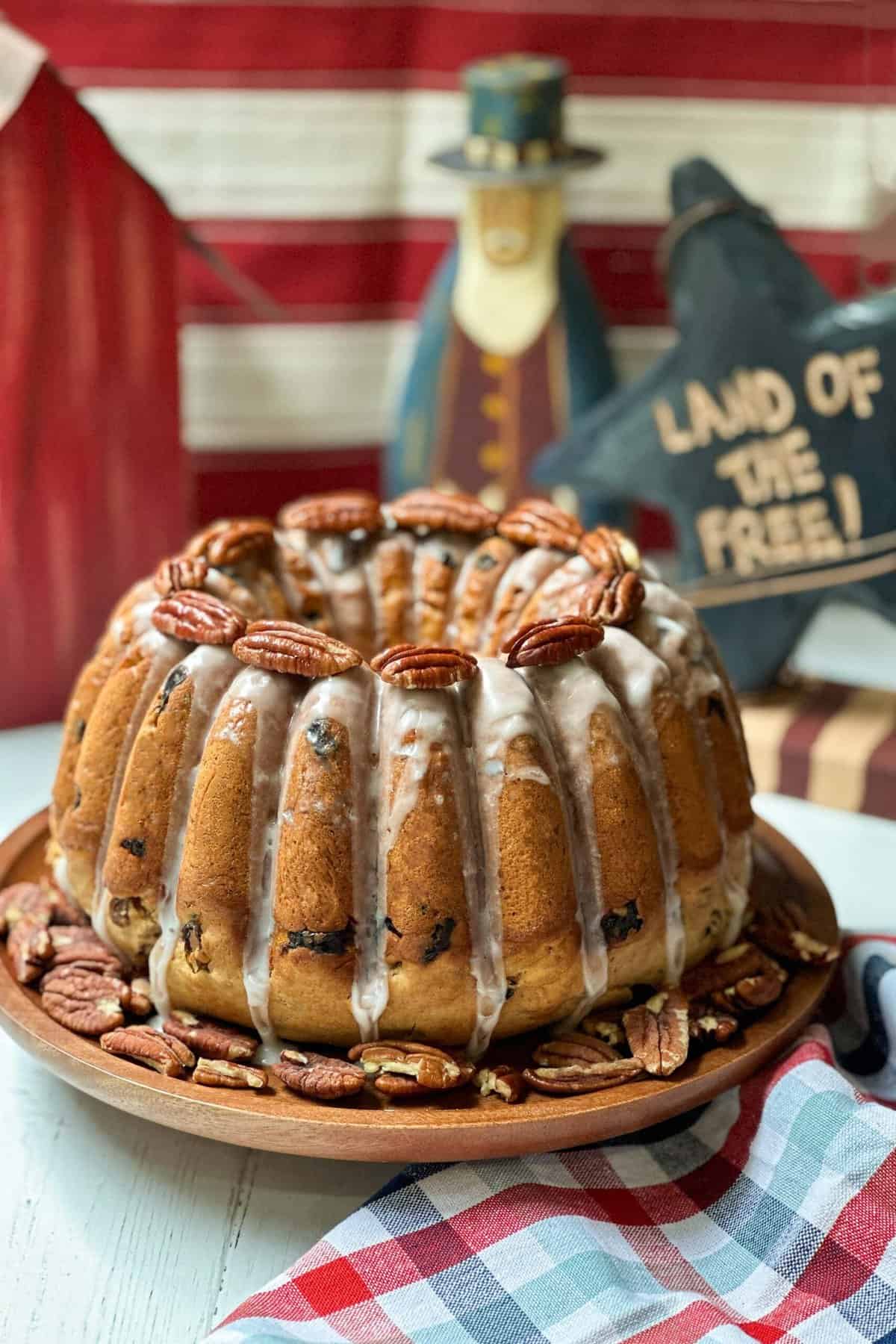 A bundt cake with a white glaze drizzled over it sits on a wooden plate. The cake is decorated with pecans on top and around the plate. In the background, there is a patriotic decoration with a sign that reads "LAND OF THE FREE!" and a checked cloth is draped beside the plate.