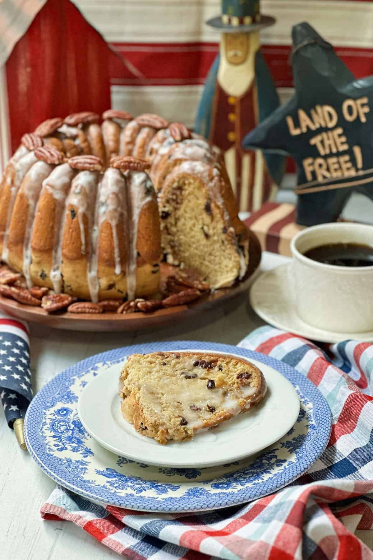 A slice of Election cake on a blue and white plate sits on a red, white, and blue checkered cloth next to a white cup of coffee. The whole bundt cake, drizzled with icing and decorated with pecans, is in the background. A sign reads "LAND OF THE FREE!".