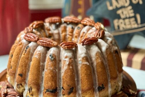 A Bundt cake topped with glaze and pecans is displayed on a checkered cloth. A sign behind it reads "Election Cake: Taste of 18th-Century History" with festive stars and a flag design. A smaller sign in the background says "LAND OF THE FREE!" Fireworks graphic in the corner, celebrating the timeless election cake.