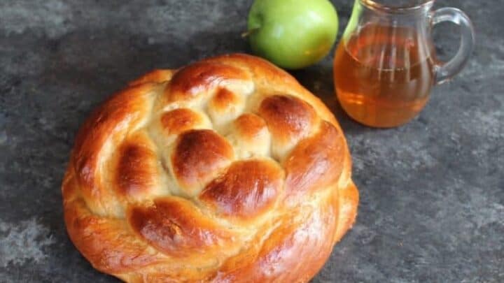 A round braided loaf of challah bread sits on a dark surface alongside a green apple and a clear glass pitcher of honey.