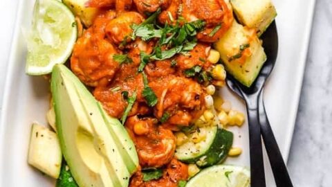A white plate filled with a colorful vegetable salad, including zucchini, corn, avocado slices, lime wedges, and cilantro. The salad is topped with a rich red sauce. Two black utensils are placed on the plate, ready to serve.