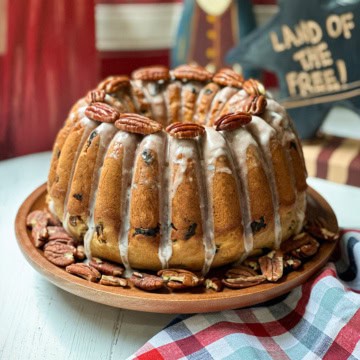 An election cake with white icing drips, studded with pecans and raisins, sits on a wooden plate surrounded by more pecans; Americana decor and a plaid cloth are in the background.