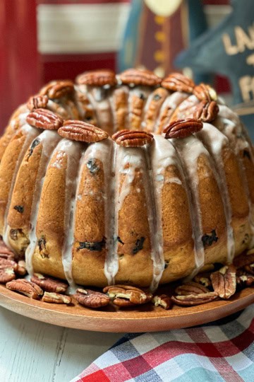 An election cake with white icing drips, studded with pecans and raisins, sits on a wooden plate surrounded by more pecans; Americana decor and a plaid cloth are in the background.