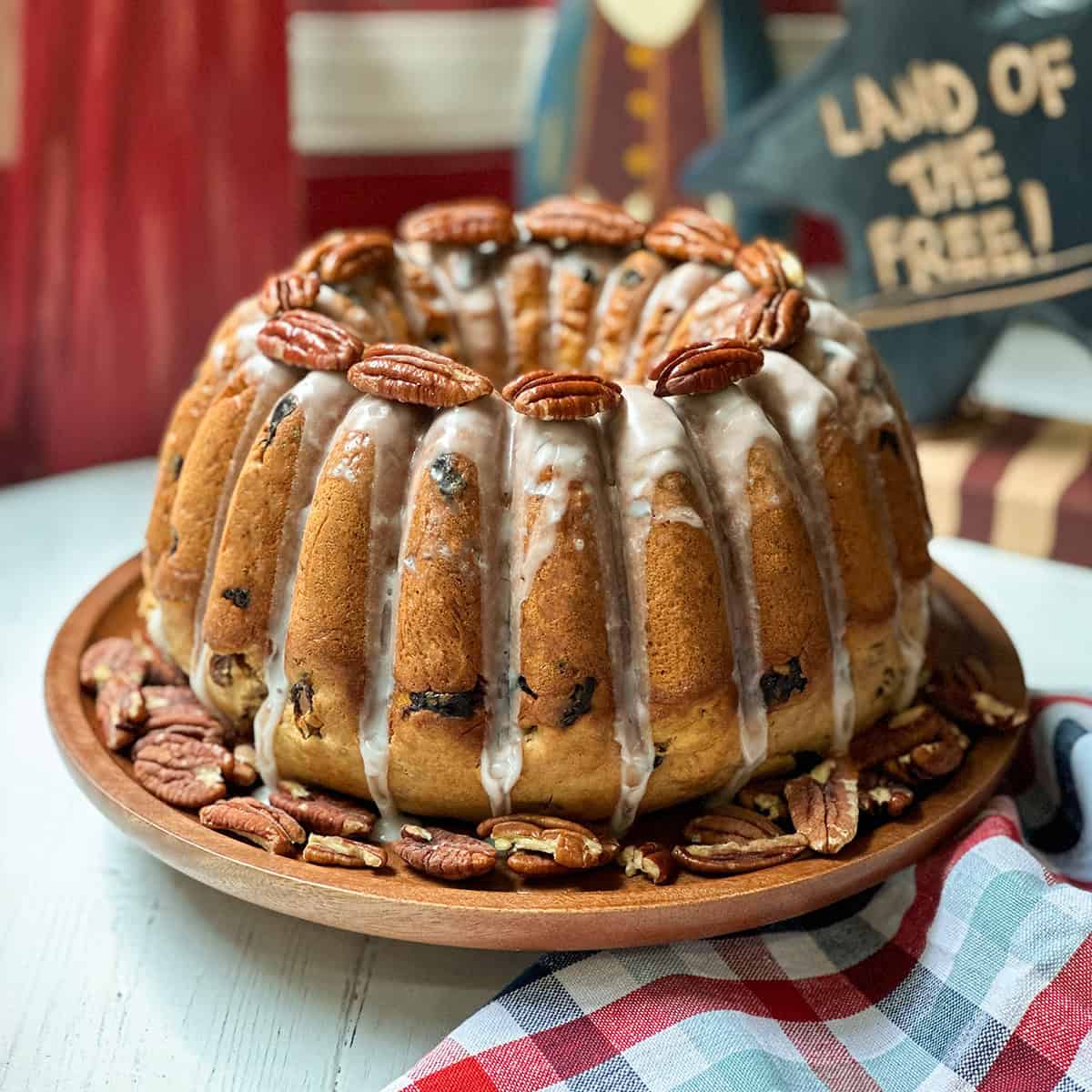 An election cake with white icing drips, studded with pecans and raisins, sits on a wooden plate surrounded by more pecans; Americana decor and a plaid cloth are in the background.