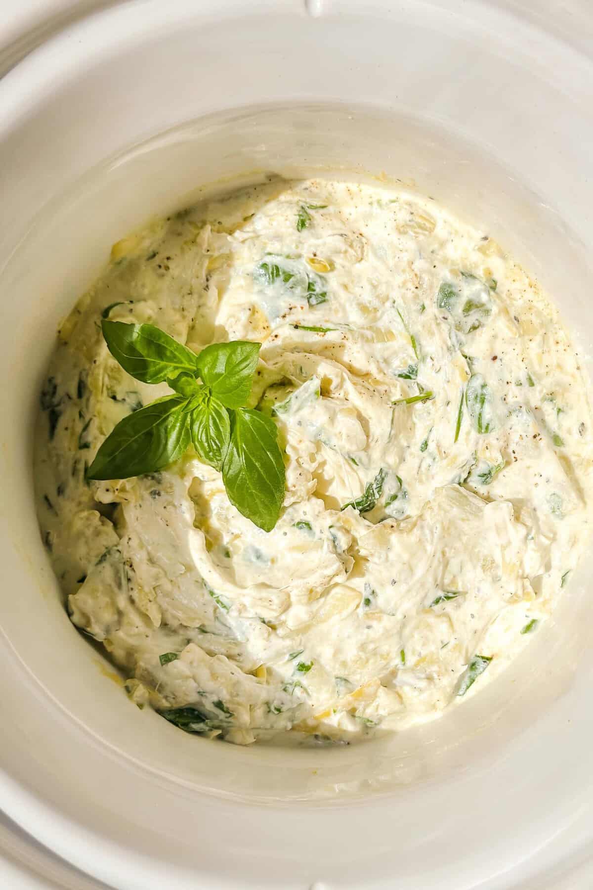 Top closeup view of Spinach Artichoke Dip in a white crockpot insert with fresh basil.