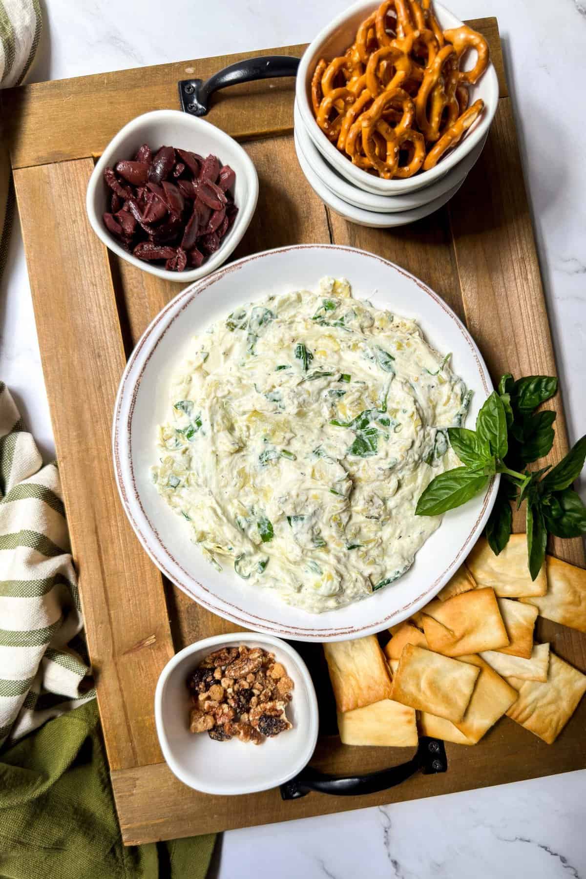 Top view of Spinach Artichoke Dip in a white bowl on a wooden serving tray with pita chips, pretzels, and fresh herbs.