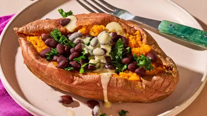 A baked sweet potato is split open and topped with black beans, kale, and a drizzle of creamy sauce. This vibrant dish, reminiscent of Mediterranean flavors, is elegantly served with a green-handled fork on a pink napkin. In the background, you'll spot a bowl of sauce and a bottle of oil.
