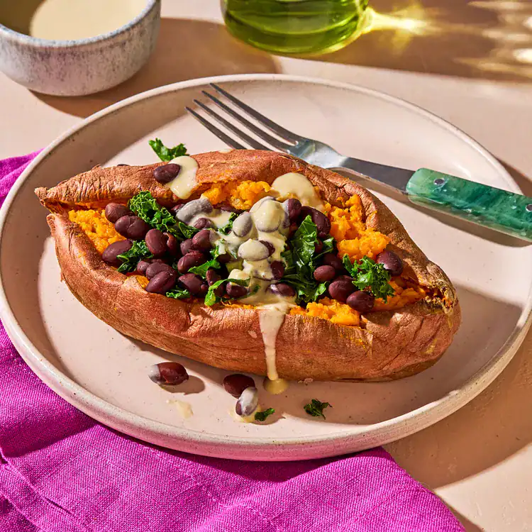 A baked sweet potato is split open and topped with black beans, kale, and a drizzle of creamy sauce. This vibrant dish, reminiscent of Mediterranean flavors, is elegantly served with a green-handled fork on a pink napkin. In the background, you'll spot a bowl of sauce and a bottle of oil.