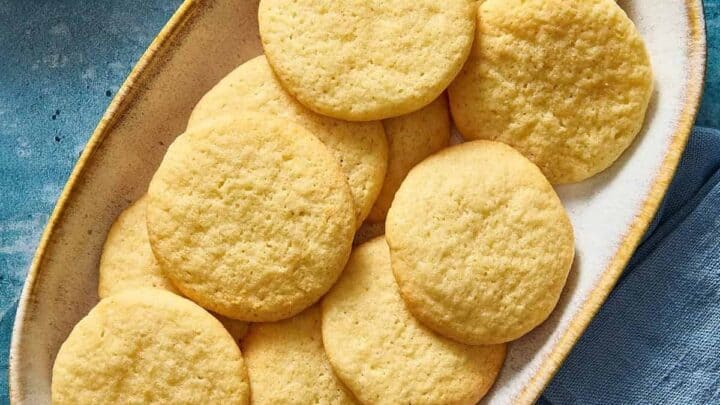 An oval plate filled with several golden-yellow cookies is placed on a blue surface. Another small plate with a single cookie is partially visible in the upper left corner. A folded blue napkin is present in the lower right corner.