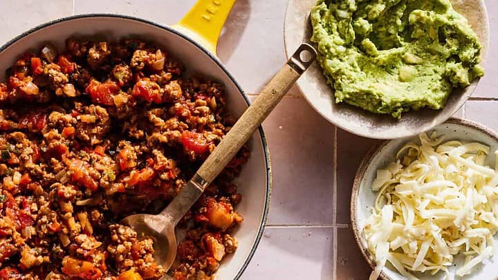 Overhead view of a meal setup including a skillet with seasoned ground meat and tomatoes, a bowl of guacamole, a bowl of shredded cheese, and a plated burger topped with the meat mixture, cheese, and guacamole for a high protein Mediterranean diet dinner. A yellow cloth napkin lies to the left.