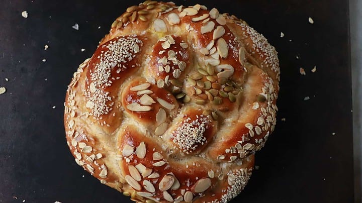 A close-up of a round, braided bread on a dark surface. The bread is golden-brown and topped with a mix of sesame seeds, almond slices, and pumpkin seeds. The surface shows a glossy finish, indicating it has been well-baked.