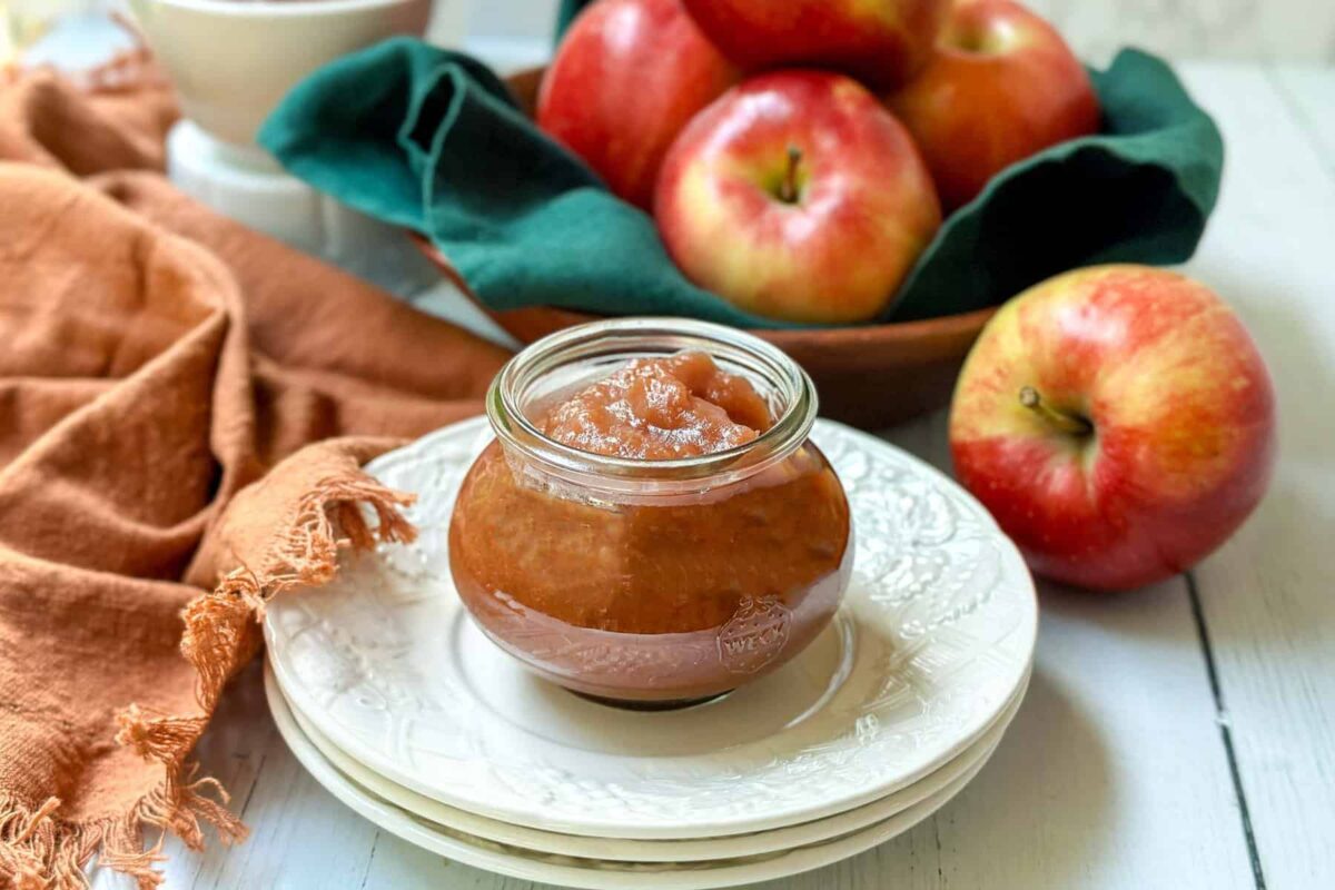 Horizontal side view of Instant Pot apple butter in a glass jar in front of a wooden bowl filled with apples.