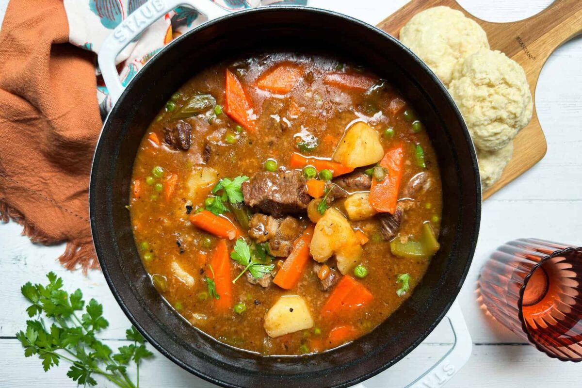 A hearty stew filled with chunks of beef, carrots, potatoes, peas, and celery in a rich broth is served in a black bowl. The bowl is accompanied by a wooden board with two biscuits, a bunch of fresh parsley, an orange cloth napkin, and a candle holder.