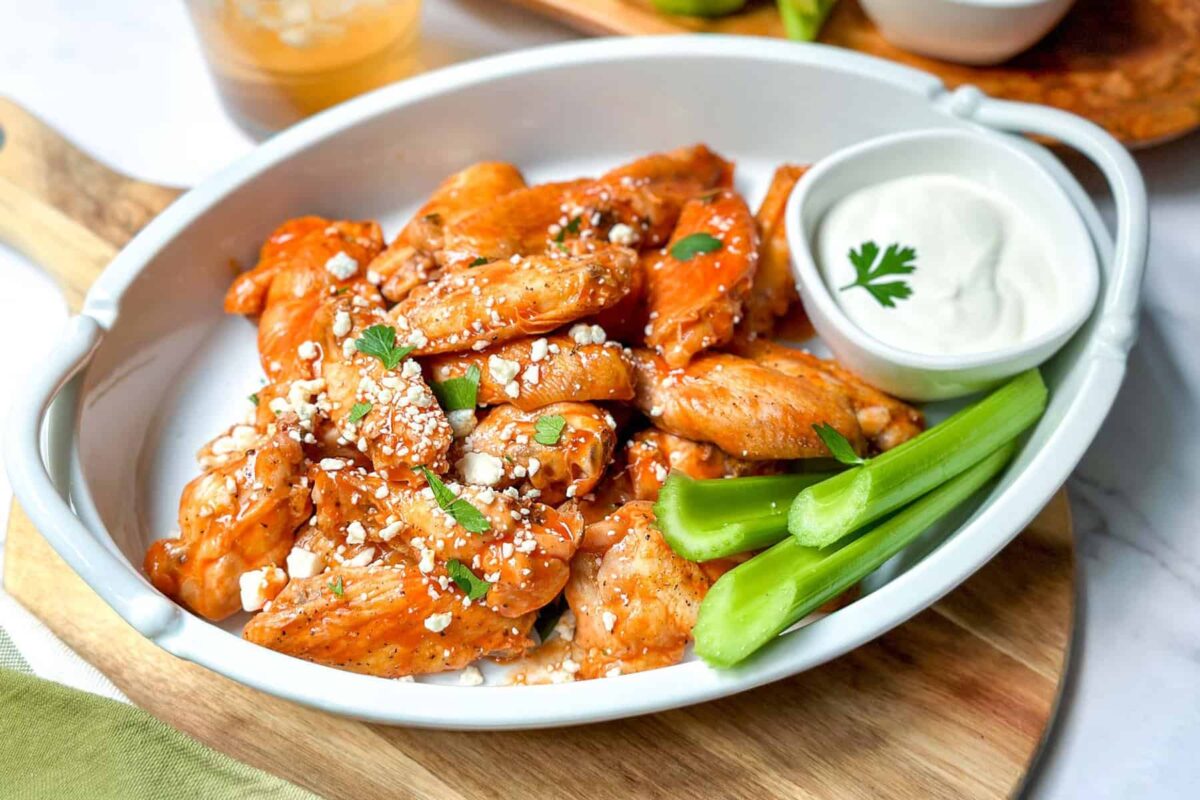 Side horizontal view of baked chicken wings in a white serving platter with buffalo sauce, dipping sauce and celery sticks for serving.