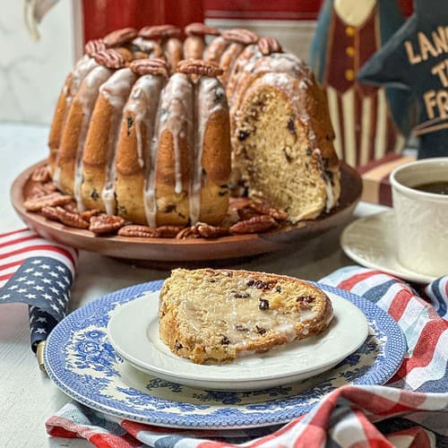 A bundt cake with icing and pecans on a plate, a slice cut and served on a blue and white plate, next to an American flag, a cup of coffee, and a red, white, and blue cloth. A sign in the background reads "LAND OF THE FREE.