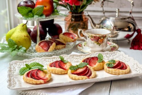 A delicate tea setting features a plate of plum tart garnished with fresh mint, alongside a china teacup, a silver teapot, and a crystal fruit stand with pears and plums. A vase with red flowers adds a touch of elegance to the background.