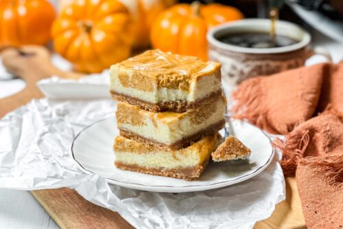 A plate holds a stack of three pumpkin cheesecake bars with a bite taken out of the top one. The dessert is layered with a crumbly crust, creamy filling, and pumpkin topping. In the background, there are mini pumpkins, a cup of coffee, and an orange napkin.