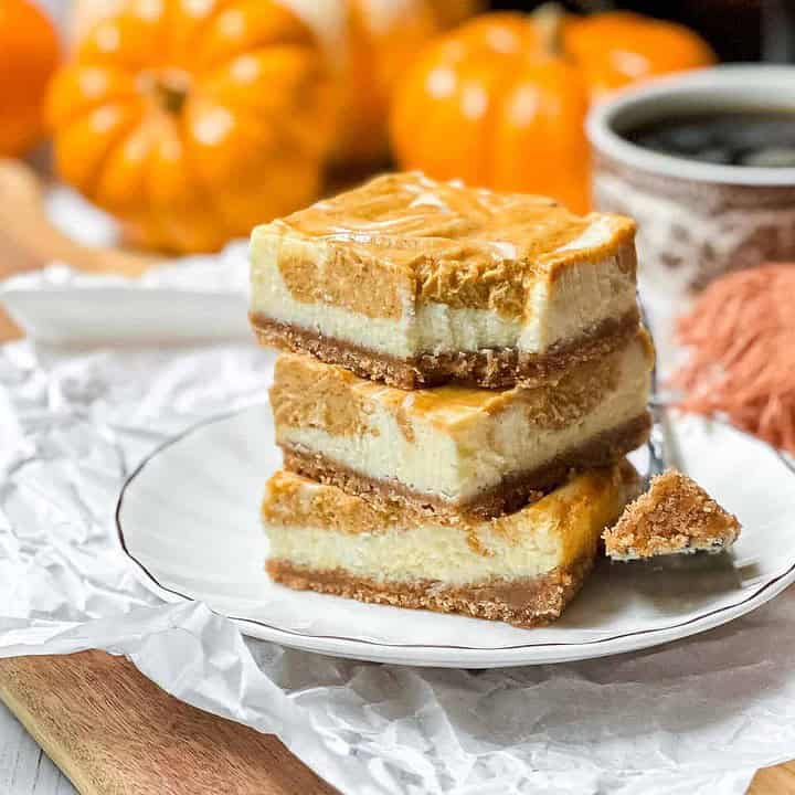 A plate holds a stack of three pumpkin cheesecake bars with a bite taken out of the top one. The dessert is layered with a crumbly crust, creamy filling, and pumpkin topping. In the background, there are mini pumpkins, a cup of coffee, and an orange napkin.