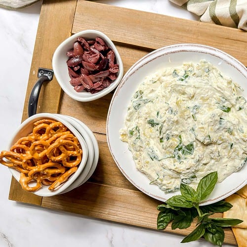 Top view of Spinach Artichoke Dip on a wooden serving board with toasted pita chips, pretzels, nuts & olives.