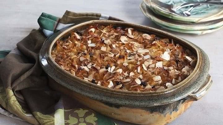 A ceramic baking dish filled with a baked casserole topped with crispy onions and almonds. The dish is placed on a checkered brown and green cloth, with a stack of plates and forks in the background.