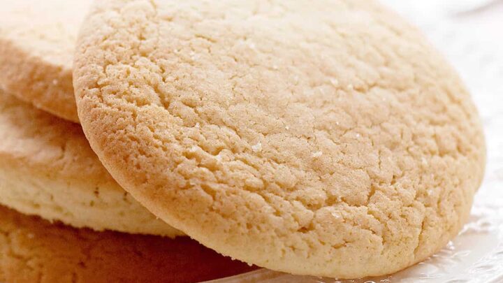 Close-up of a plate with three large, round sugar cookies stacked slightly on top of each other, resembling classic teacakes. The cookies have a golden-brown, slightly cracked surface and appear soft and chewy. The white plate and bright background suggest a clean, inviting setting.