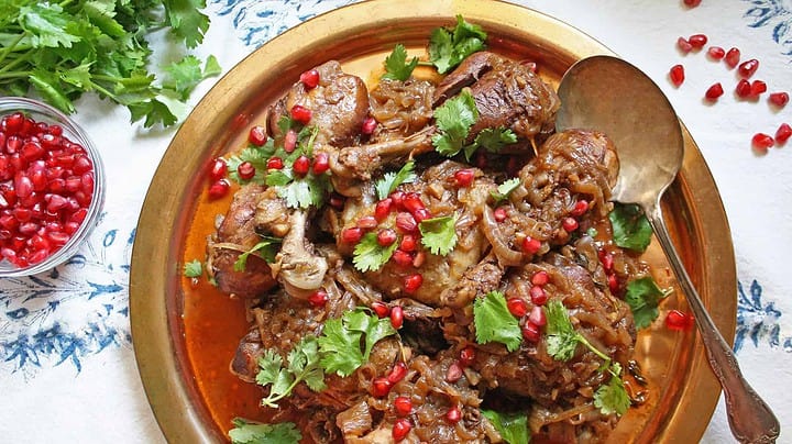 A brass plate holds a serving of cooked meat pieces topped with caramelized onions, fresh cilantro, and pomegranate seeds. A spoon is placed to the side on the plate. Pomegranate seeds and cilantro are scattered on the white and blue tablecloth around the plate.