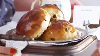A person holding a metal tray with three shiny, freshly baked raisin teacakes. The background includes a white mug and a small, folded card with text partially visible. The person has red nail polish and holds the tray with a cloth.