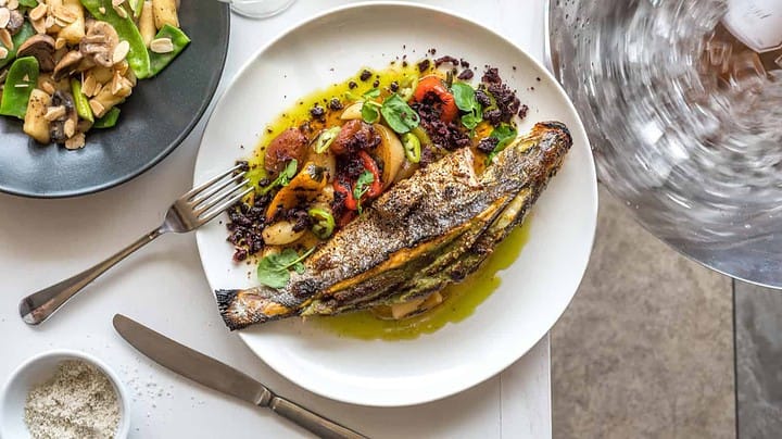 A beautifully plated dish of grilled fish with colorful vegetables and herbs sits on a white plate, accompanied by a glass of white wine. Another plate of mixed greens and mushrooms is nearby, and a bowl of salt is on the side. A person is pouring water into a glass.