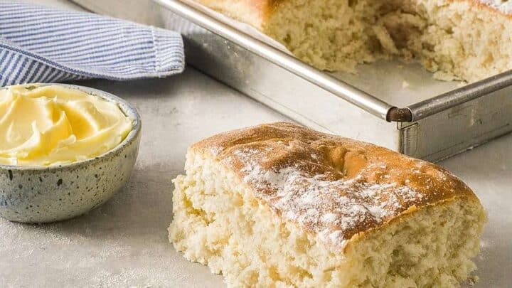 A tray of freshly baked focaccia bread with a piece cut out and placed in the foreground. The bread has a golden crust and is lightly dusted with flour. A small bowl of butter and a blue-striped cloth are next to the tray on a light gray surface.