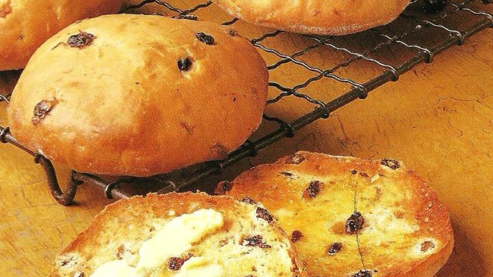 Freshly baked bread rolls with raisins cooling on a wire rack. One roll is sliced open, with butter spread on one half. A small dish of butter is placed near the sliced roll. The background is a warm, rustic wooden surface.
