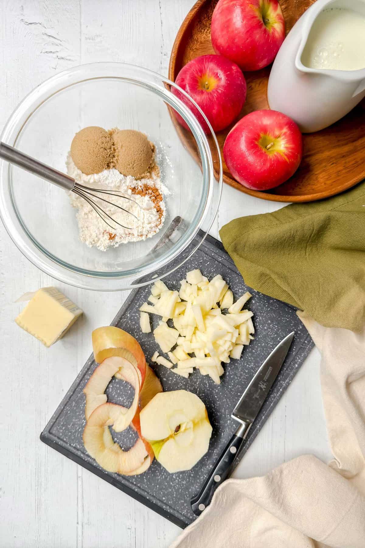 Top-down view of baking ingredients including a bowl with flour, brown sugar, and spices, a cutting board with chopped apples, apple slices, a knife, a block of butter for an apple mug cake, three whole apples on a plate, and a pitcher of milk on a white wooden surface.