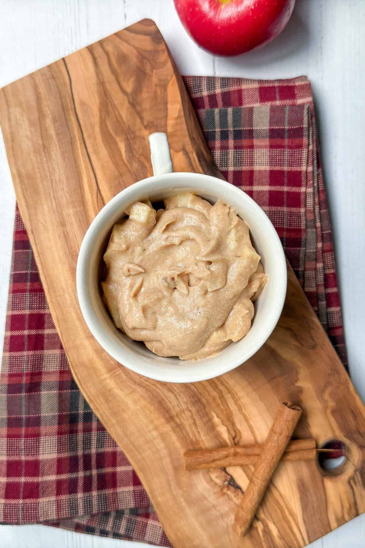 A mug of unbaked apple mug cake batter with chunks of apple sits on a wooden board placed atop a red and brown checkered cloth. Next to the cup are two cinnamon sticks. An apple is partially visible at the top right corner of the image.