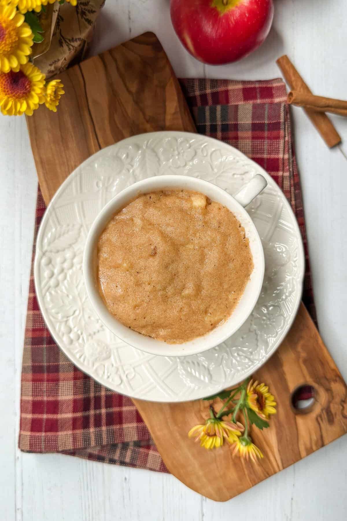 A white mug filled with just baked apple cake rests on a matching saucer atop a red plaid cloth and wooden board. Surrounding the mug are yellow flowers, a red apple, and two cinnamon sticks arranged in a cross. The setup is on a light-colored wooden table.